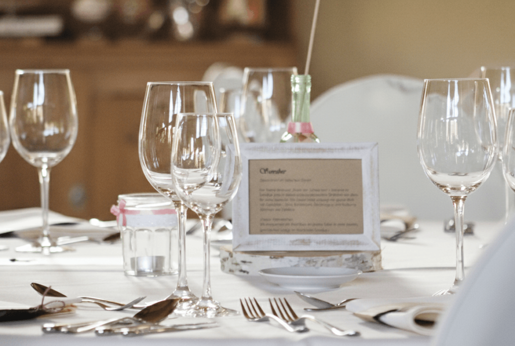 Eine festlich gedeckte Tafel in einem hellen Raum. Im Fokus steht ein kleiner weißer Bilderrahmen mit einer Menükarte auf Kraftpapier, flankiert von eleganten Weingläsern und feinem Silberbesteck auf einer weißen Tischdecke. Im Hintergrund