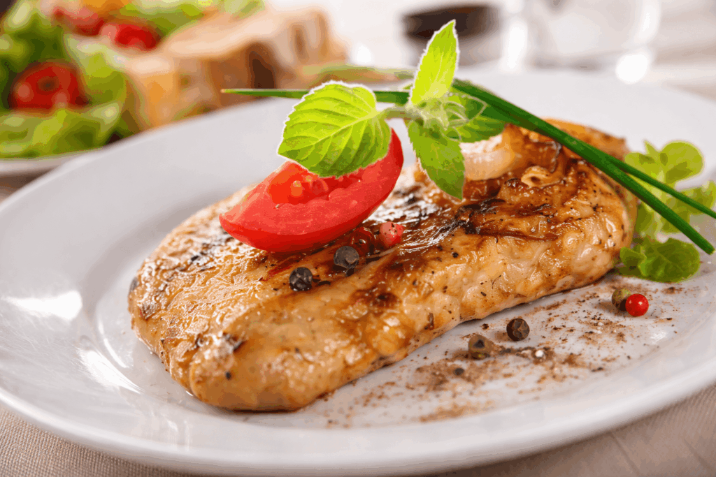 Gegrilltes Hähnchenbrustfilet auf weißem Teller, goldbraun gebraten, garniert mit einer Tomatenscheibe, frischer Minze und buntem Pfeffer, im Hintergrund leicht unscharfer Salat und Brot.